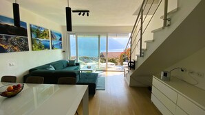 Ein modernes Wohnzimmer mit einer Couch, einem Esstisch mit Obstschale und Meerblick durch große Fenster.