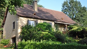 Een stenen huis met schuin dak, omgeven door weelderige begroeiing en bomen.