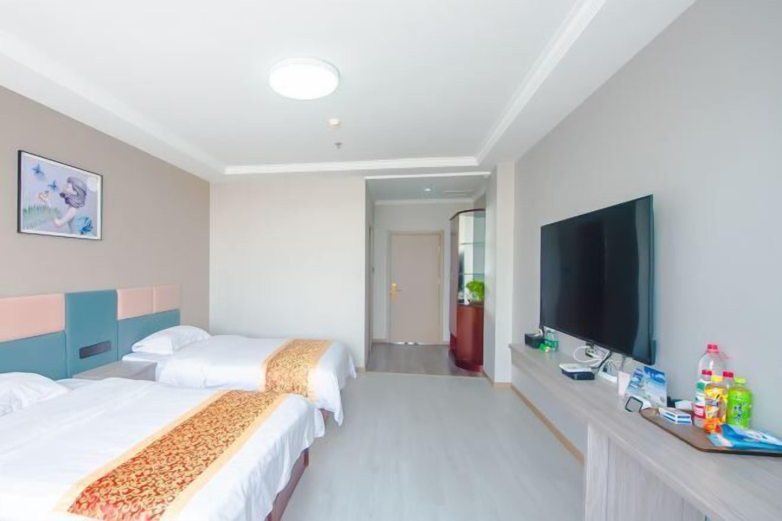 Ein Hotelzimmer mit zwei Betten, einem Fernseher, einem Schreibtisch mit diversen Gegenständen und einem Gang zu einem weiteren Zimmer.