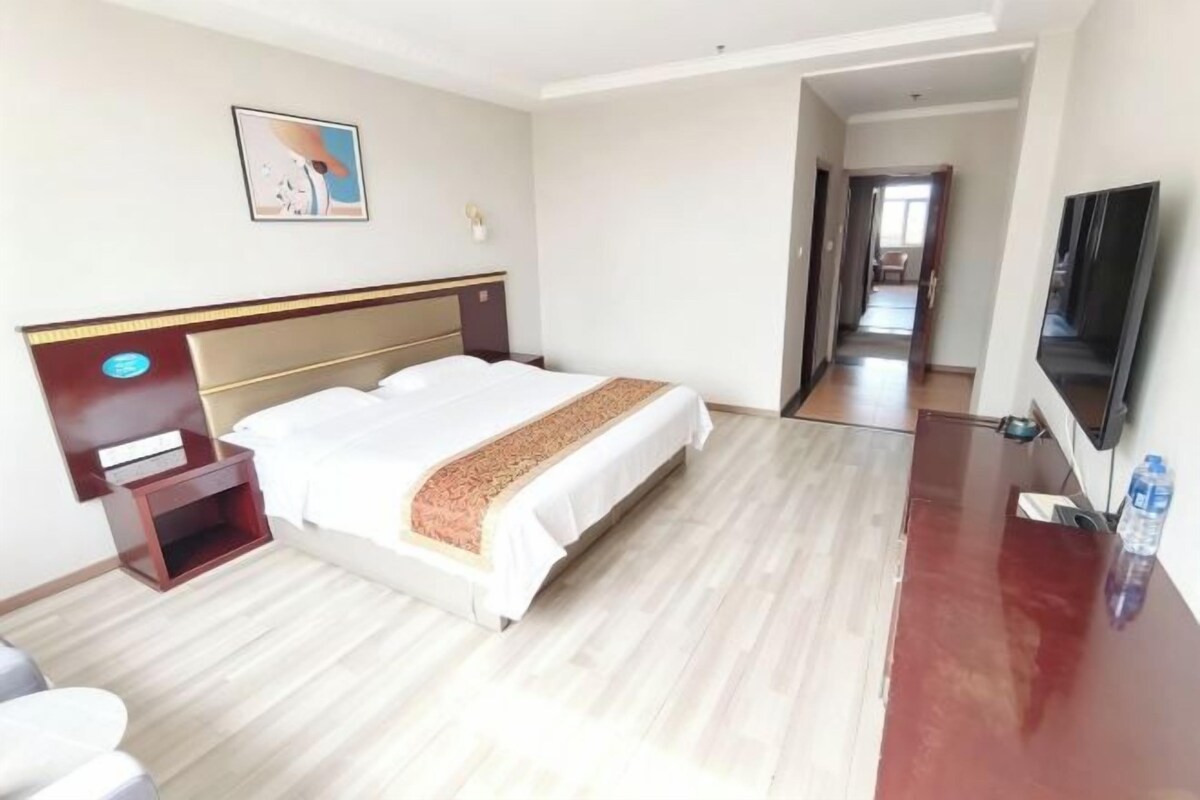 A hotel room with a bed, a desk, a chair, a TV, and a door leading to another room.