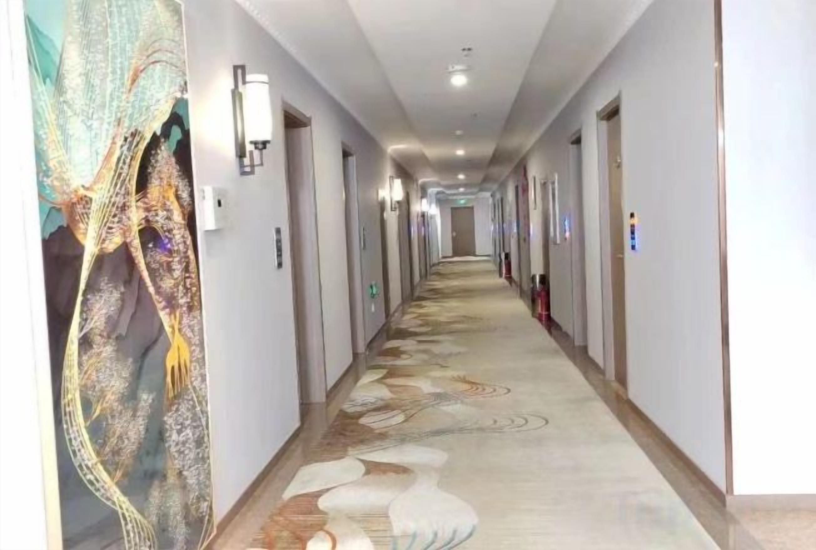A hotel corridor with a patterned carpet, doors on both sides, and wall-mounted light fixtures.