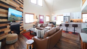 A modern living room with a fireplace, a sofa, a TV, and a kitchen area with bar stools. - Mountain Modern Family Home near Grand Targhee - Hot Tub (Driggs)