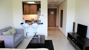 A modern living room with a sofa, a coffee table, a TV stand, and a kitchen area in the background. - KSLV - Furnished Studio (Dubai)