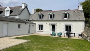 Une maison à deux étages, avec une façade blanche, un toit en tuiles et un petit jardin à l’arrière, comprenant une table et des chaises.