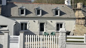 Une maison à deux étages, avec une clôture en bois peinte en blanc, un mur de pierres et un petit jardin agrémenté de chaises vertes.