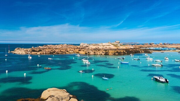Une zone côtière où des bateaux sont amarrés dans des eaux turquoise et limpides, avec un paysage rocheux et un ciel dégagé.
