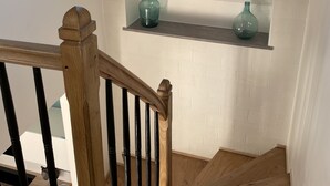 Un escalier en bois, surmonté d’une lucarne, mène à un mur de briques blanches où sont disposées deux étagères sur lesquelles reposent des bouteilles en verre vert.