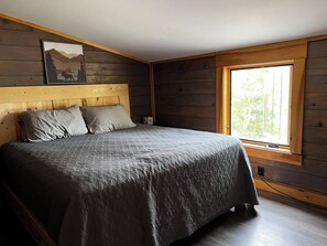 A bedroom with a wooden headboard, a large bed with grey bedding, a window with a view of trees, and a painting on the wall. - Cozy Modern Cabin (Rockwood)