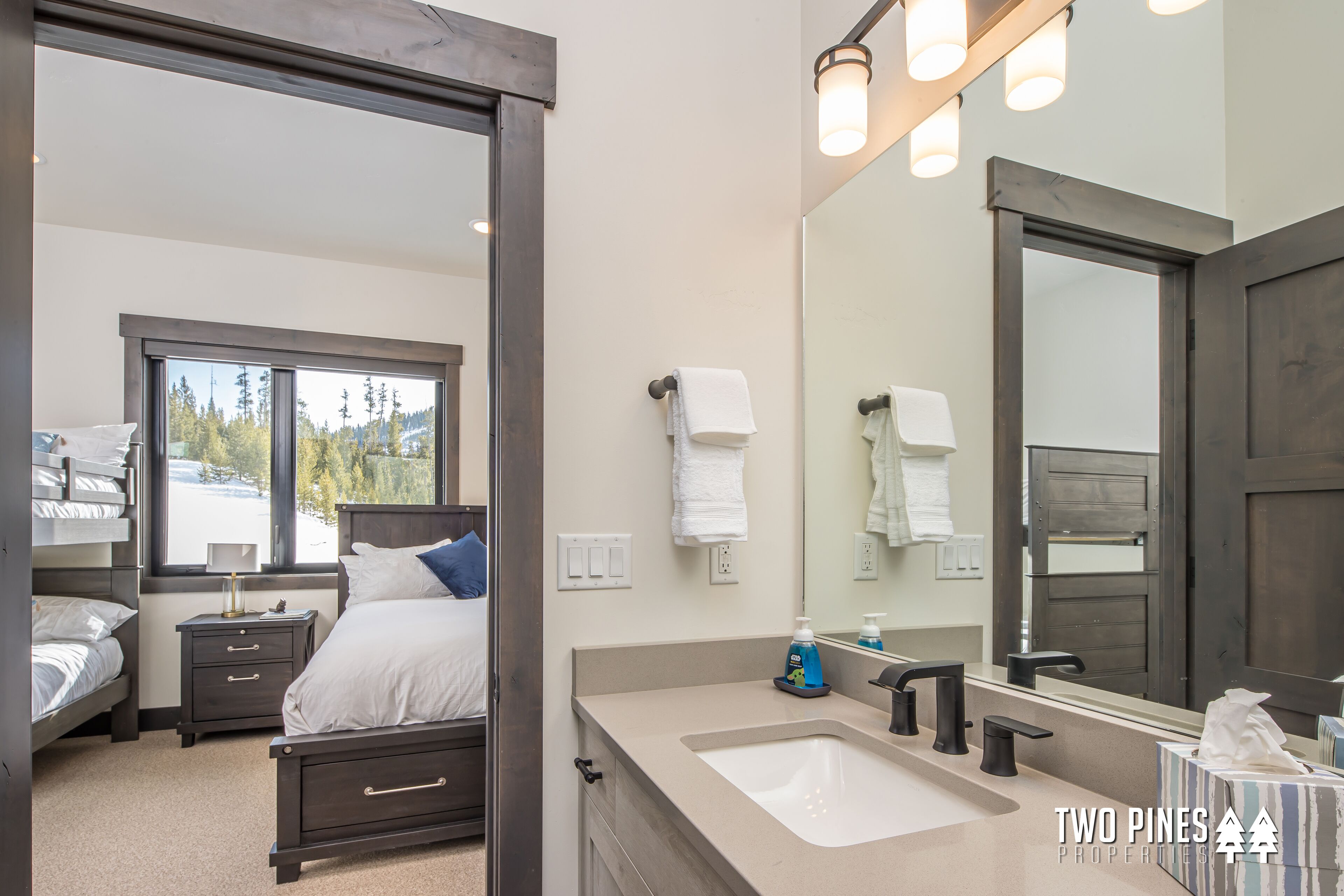 A modern bedroom with a large bed, bedside tables, a dresser, a window with a view of trees, and a bathroom with a sink, mirror, and towel rack.