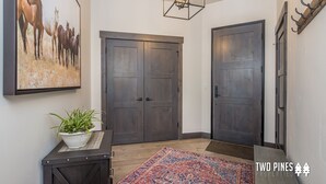 Un couloir avec deux portes en bois sombre, un grand tableau représentant un cheval, un luminaire suspendu et une plante en pot posée sur une petite table.
