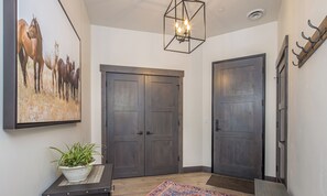 Un couloir avec deux portes en bois sombre, un grand tableau représentant un cheval, un luminaire suspendu et une plante en pot posée sur une petite table.