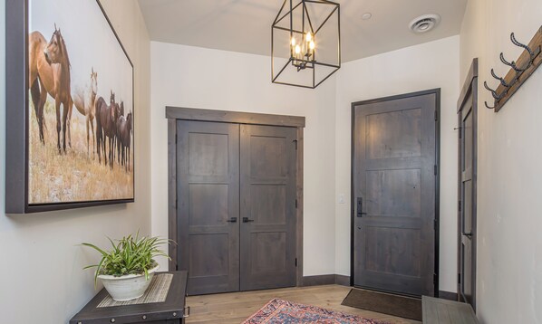 Un couloir avec deux portes en bois sombre, un grand tableau représentant un cheval, un luminaire suspendu et une plante en pot posée sur une petite table.