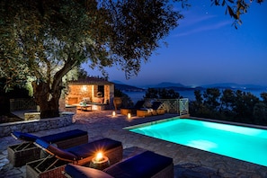 An outdoor pool area with lounge chairs, a stone wall, and a small seating area under a tree. - Villa Kalokairi comfortable and stylish stone house above Sivota (Sivota Lefkada)