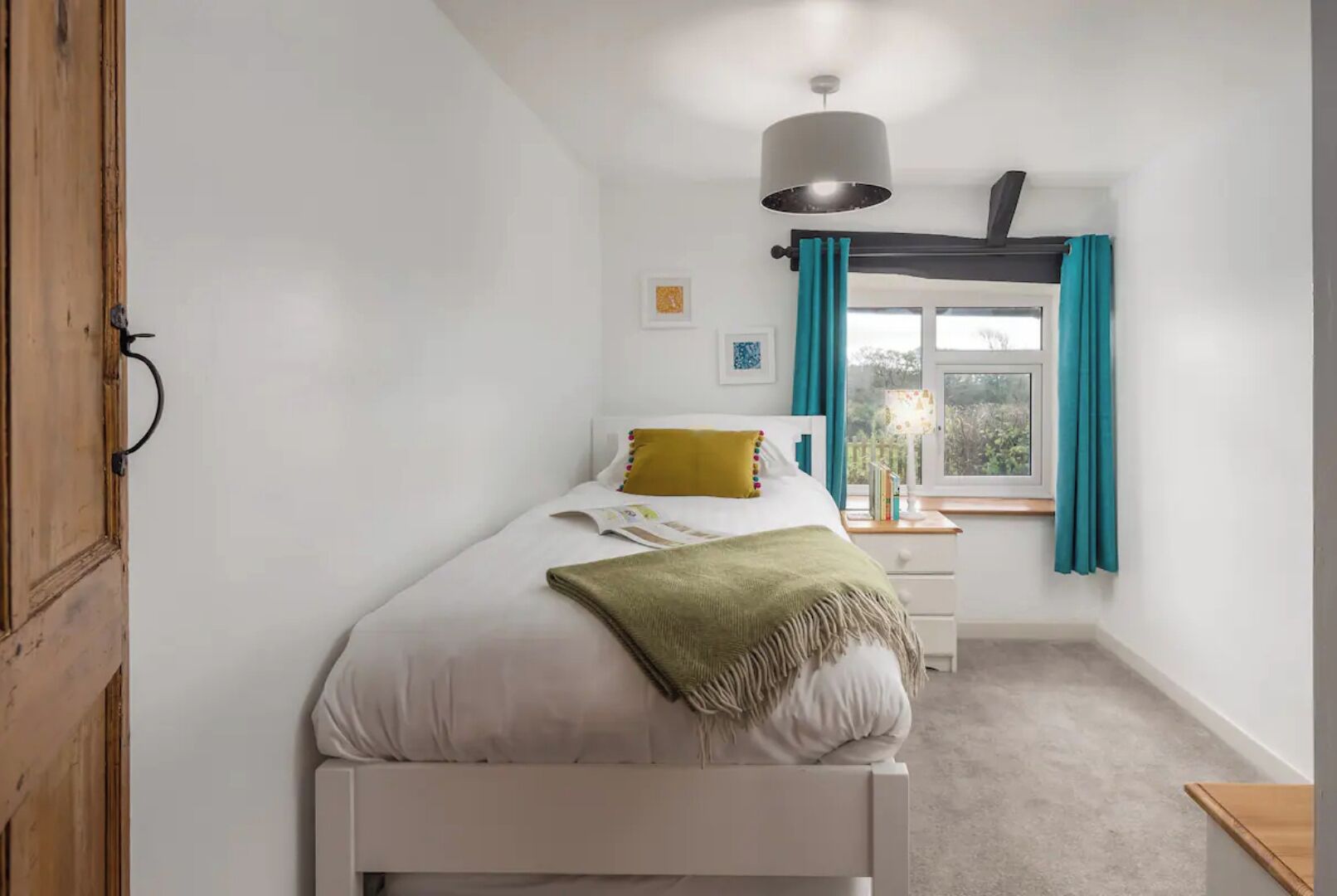 A bedroom with a bed, a bedside table, a lamp, a window with curtains, and a door.
