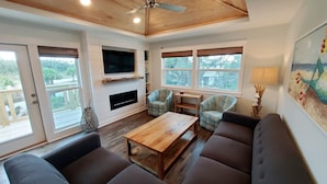A spacious living room with a wooden ceiling, a flat-screen TV mounted on the wall, a fireplace, and large windows offering views of the outdoors. - Private Pool! Easy Beach Access! (St. George Island)