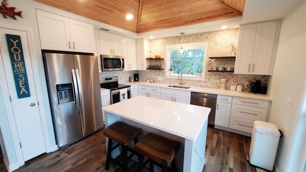 A modern kitchen with white cabinets, stainless steel appliances, a wooden ceiling, and a large island with stools. - Private Pool! Easy Beach Access! (St. George Island)