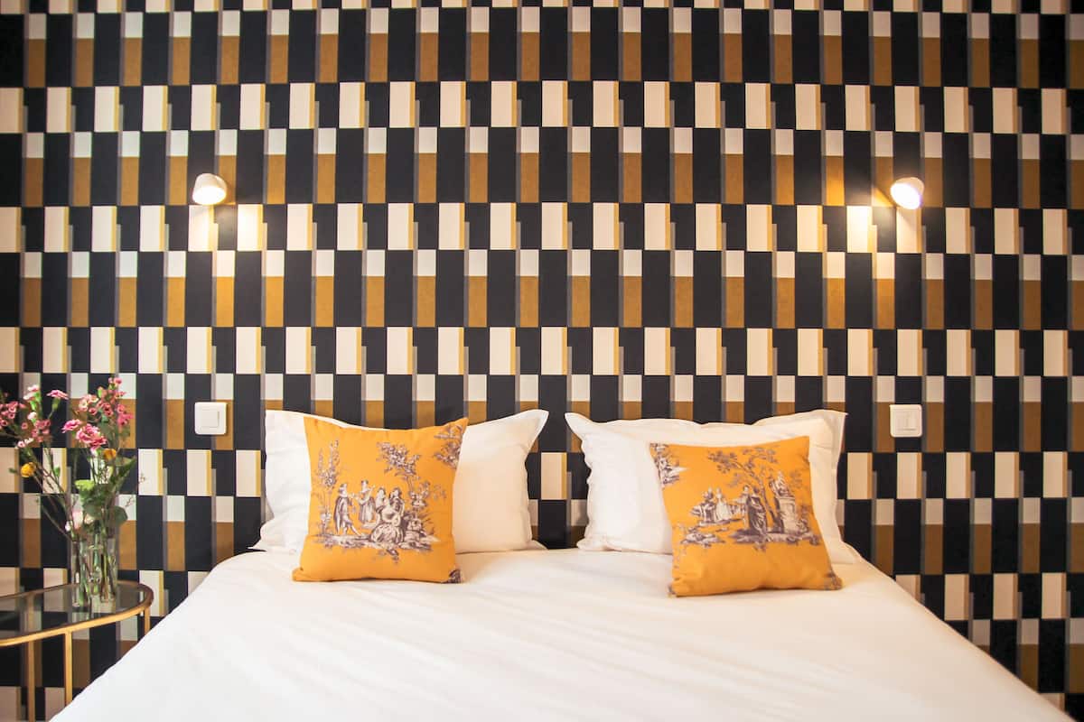 Quarto de hotel com padrão de parede xadrez, cama com roupa de cama branca e duas almofadas amarelas, e uma pequena mesa com um vaso de flores.