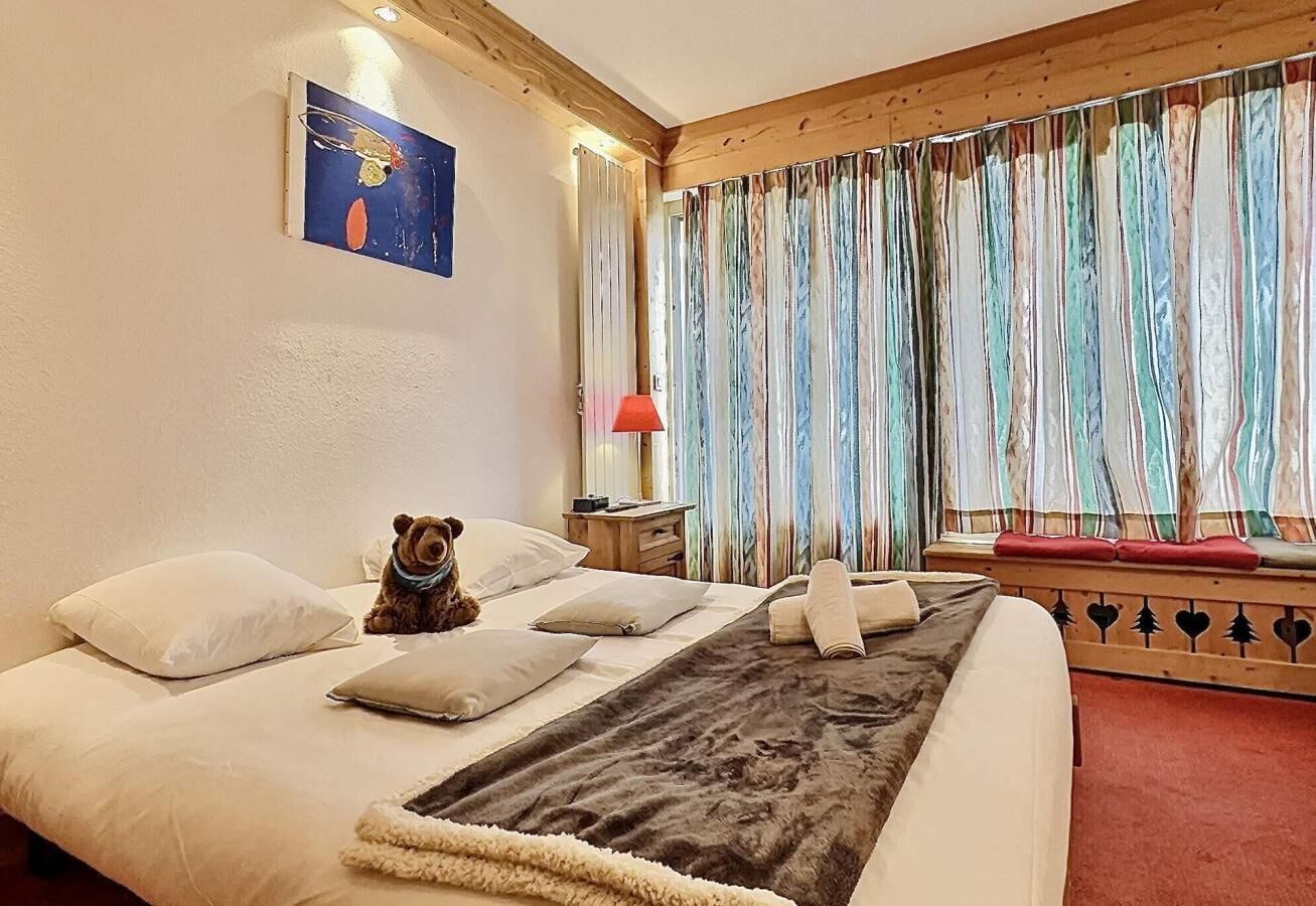 Uma cama com almofadas, um cobertor, um ursinho de pelúcia, uma mesinha de cabeceira, um abajur e uma janela com cortinas.