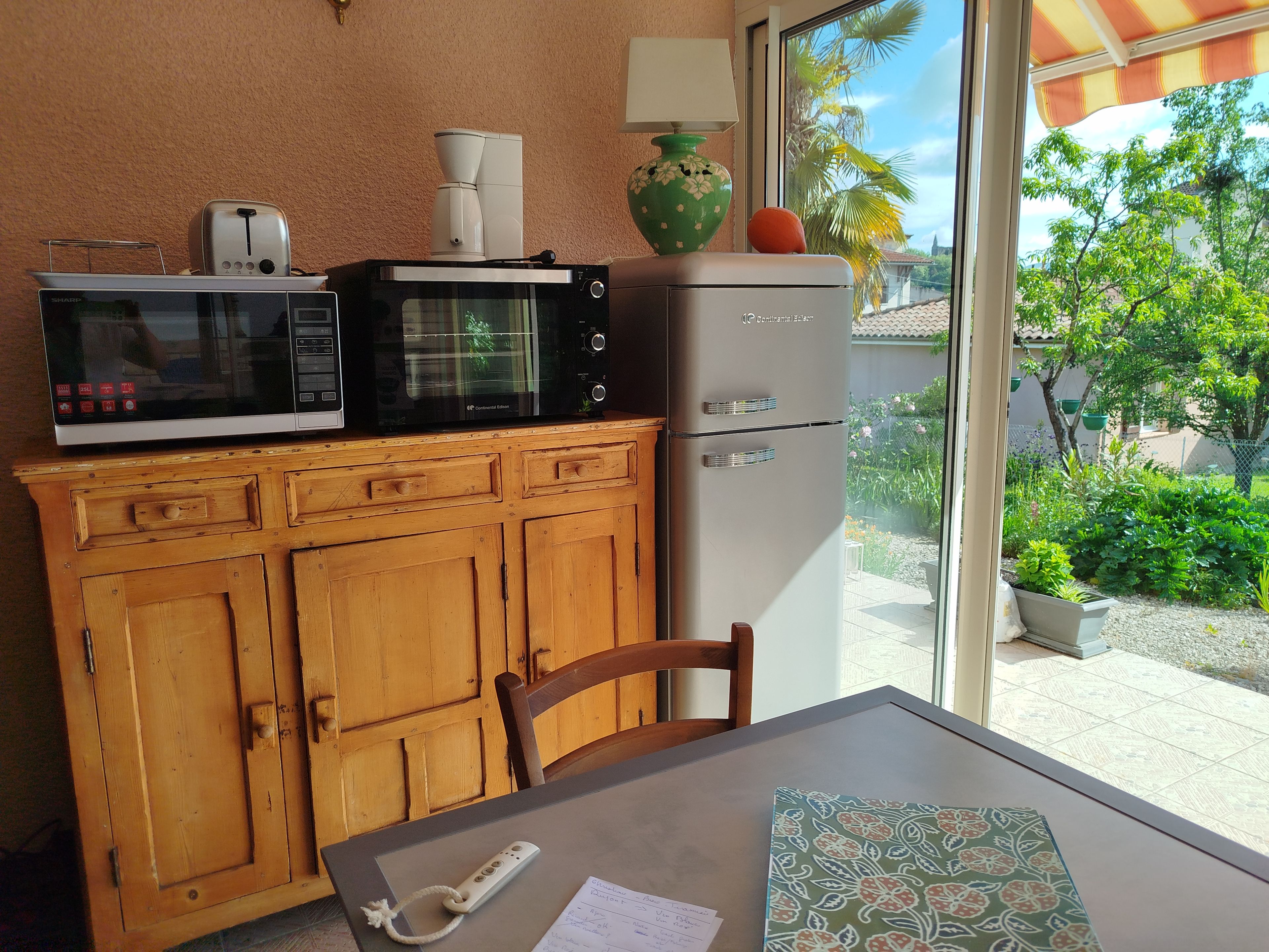Una kitchenette compacta con microondas, tostadora y refrigerador. Se ve también un mueble de madera y una mesa con un control remoto y papeles.