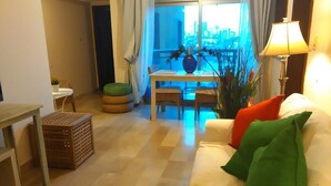 Een woonkamer met een bank, een eettafel met stoelen, een lamp en een uitzicht op een stadsbeeld door een groot raam.