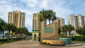 Entrada a un resort rodeado de palmeras y edificios altos, con un cartel que dice 'Beach Colony Resort'.