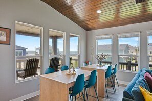 A modern dining area with a wooden table and blue chairs, large windows with views of a residential neighborhood. - The Neon Moon 3 Bedroom Home by RedAwning (Port Bolivar)