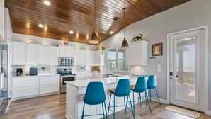 Une cuisine moderne avec des meubles blancs, un îlot central doté de tabourets de bar bleus, un plafond en bois et une grande fenêtre.