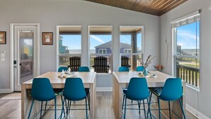 Un vaste espace de salle à manger avec une longue table et des chaises bleues, de grandes fenêtres offrant une vue sur une maison et une clôture, et un plafond en bois.