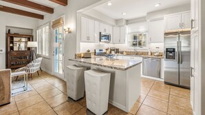 A modern kitchen with white cabinetry, a large island, stainless steel appliances, and a dining area with a white chair and table. - Luxurious 2 Story Townhome - Newly Remodeled! 3 Bedroom Condo by RedAwning (La Quinta)