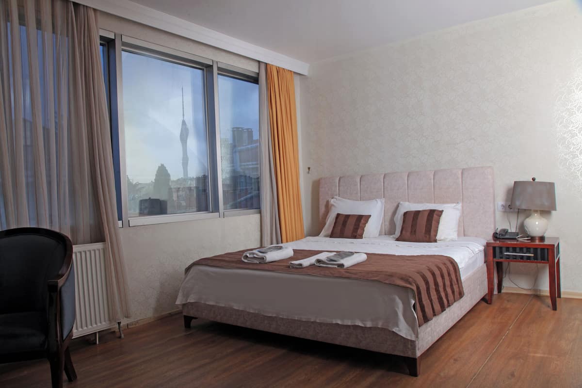 Quarto de hotel com uma cama grande, uma cadeira, mesa de cabeceira com abajur e uma janela com cortinas.