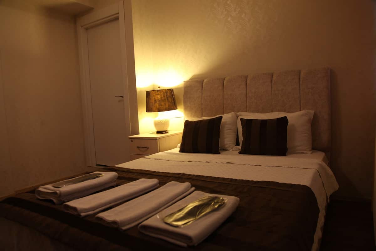 Quarto de hotel com uma cama, uma mesinha de cabeceira com abajur e toalhas dobradas na cama.