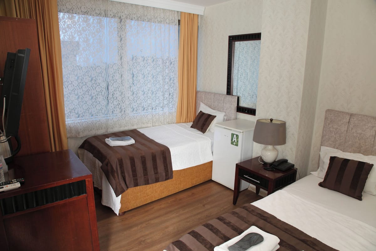 Quarto de hotel com duas camas, uma escrivaninha, uma luminária, um espelho e uma janela com cortinas.