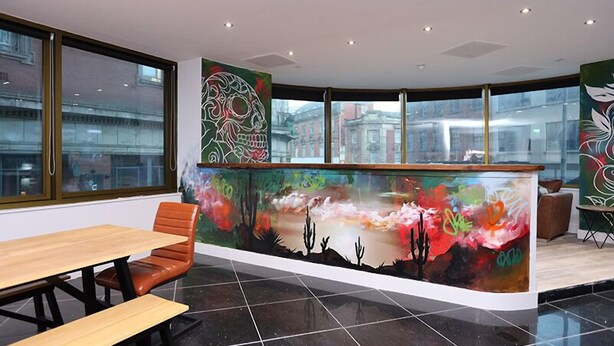 A modern reception area with a curved counter, a wooden bench, and a large window offering a view of buildings outside. - The Bling Hotel by UStay (Liverpool)