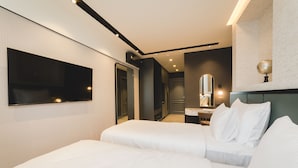 Una habitación de hotel moderna con un televisor de pantalla plana grande, dos camas con sábanas blancas, un espejo sobre la cama y un escritorio pequeño con una lámpara.
