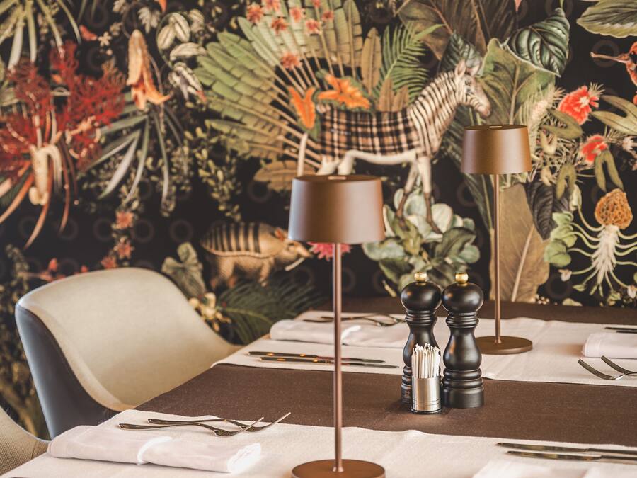 Mesa de comedor dispuesta con mantel blanco, cubiertos y dos lámparas con pantallas marrones.