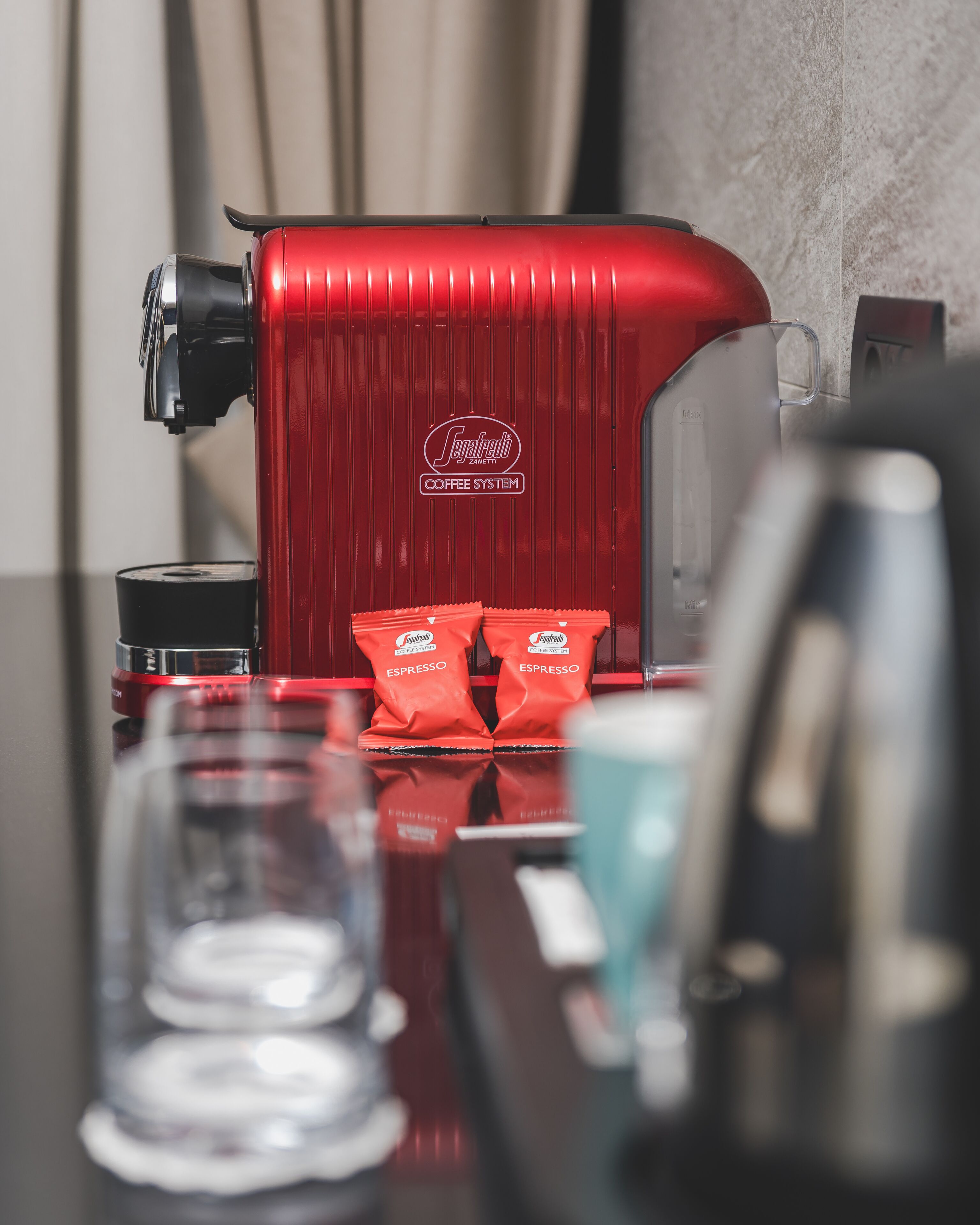 Punainen kahvinkeitin, jonka päällä pöydällä on kaksi espressokapseleita.