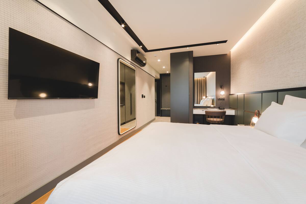 Ein modernes Hotelzimmer mit einem großen Bett, einem flachbildfernseher an der Wand, einem Schreibtisch mit Stuhl und einem Spiegel.