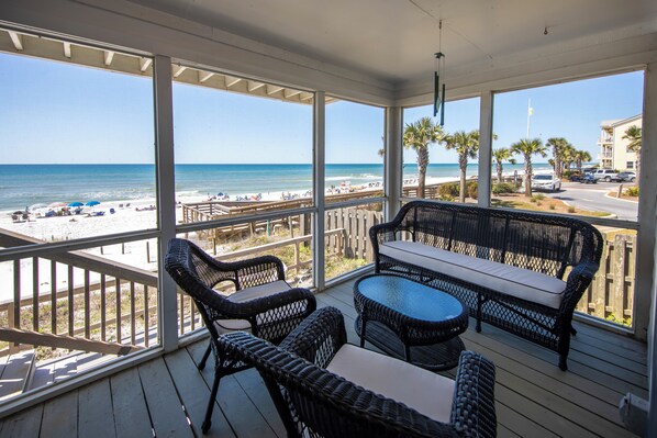 A covered outdoor seating area with wicker furniture, overlooking a beach with palm trees and a clear view of the ocean.