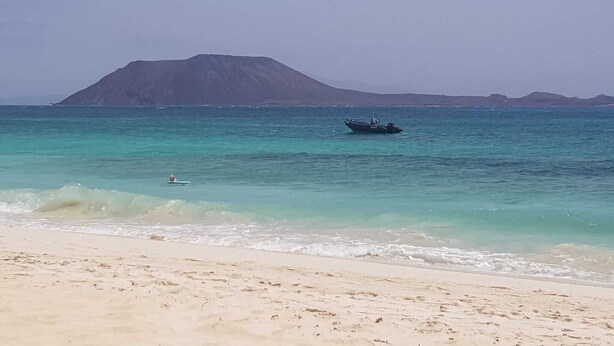 A beach with clear turquoise water, a boat in the distance, and a mountain in the background. - Oasis Royal - Beautiful Corralejo Apartment (Corralejo)