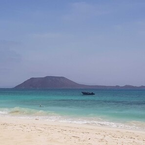 A beach with clear turquoise water, a boat in the distance, and a mountain in the background. - Oasis Royal - Beautiful Corralejo Apartment (Corralejo)