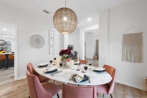 A dining area with a round table, pink chairs, a hanging light fixture, and a macrame wall hanging.