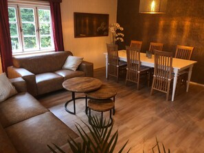 Un salon avec un canapé en cuir marron, deux petites tables en bois, une table à manger avec des chaises en bois, un vase avec des fleurs blanches et un tableau accroché au mur.