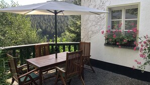 An outdoor patio with a wooden table and chairs, a large umbrella, and a window with pink flowers. - Haus Onel 1 (Schmallenberg)