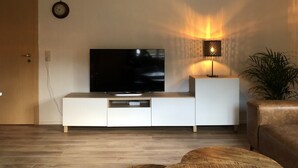 A modern living room with a flat-screen TV on a white entertainment unit, a floor lamp, and a potted plant. - Haus Onel 1 (Schmallenberg)