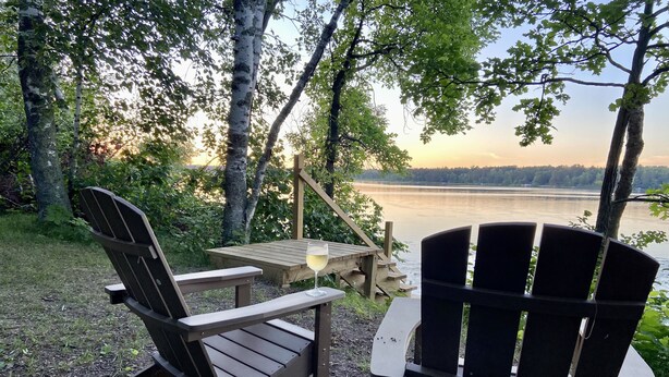 A lakeside seating area with Adirondack chairs and a table, surrounded by trees and a view of the lake.