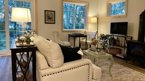 A living room with a beige sofa, a coffee table, a TV, and a fireplace. There are windows with views of trees.