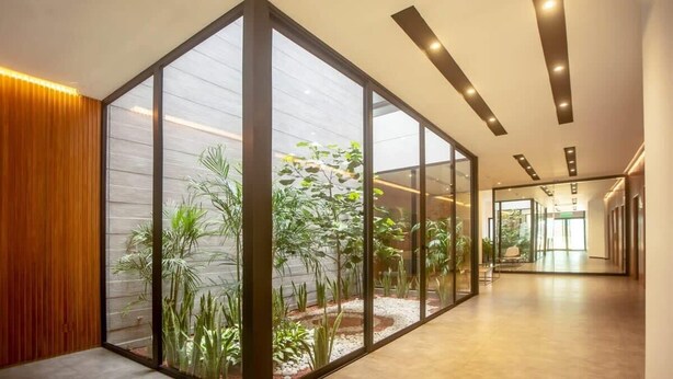 Een moderne hal met glazen wanden, een houten paneel aan de zijkant en uitzicht op een tuin vol planten.