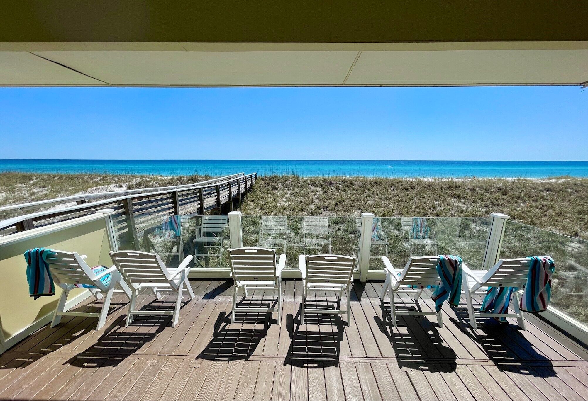 A deck with white chairs and a glass railing overlooking a beach.