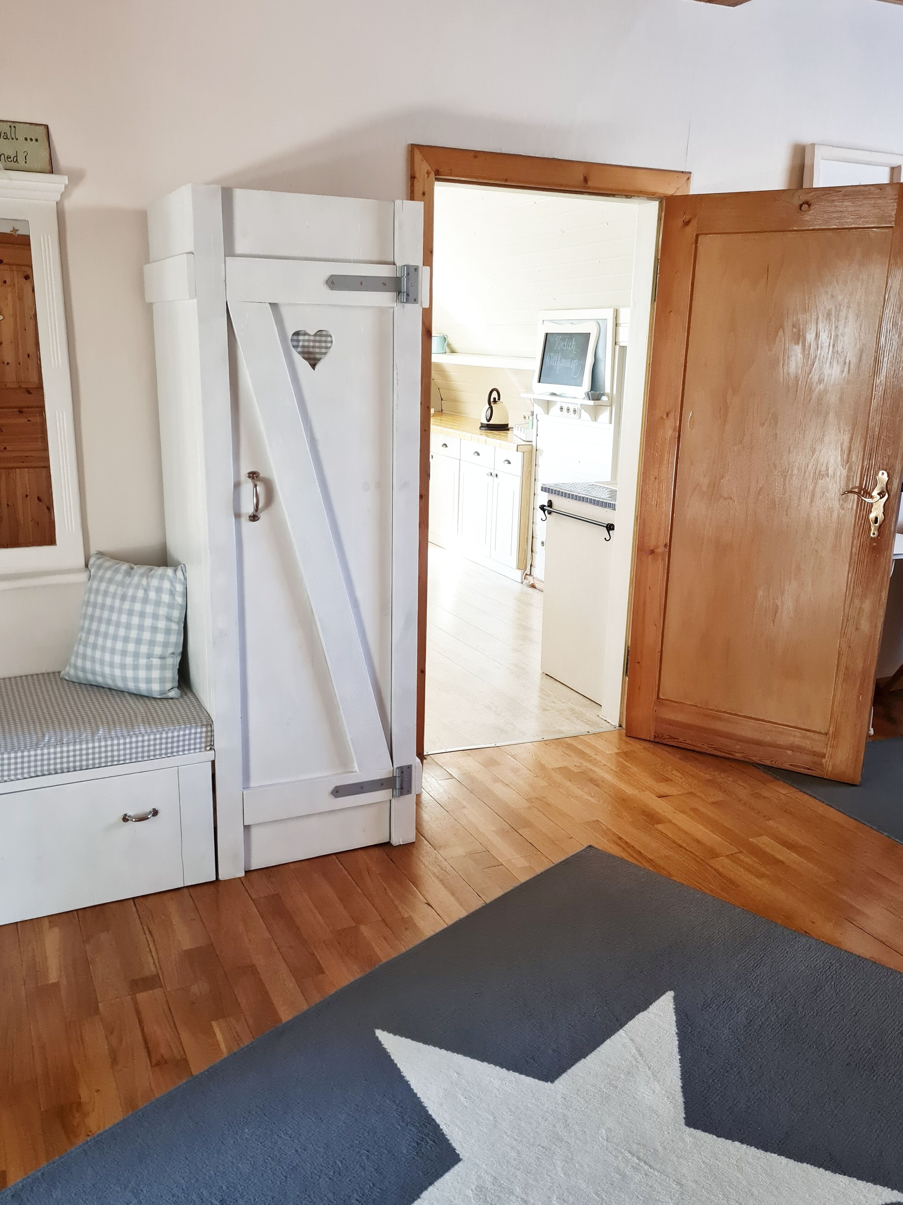 Une pièce compacte avec une kitchenette, une porte en bois, un banc doté d’un coussin à carreaux et un tapis orné de motifs en forme d’étoiles.