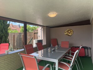 A covered patio with a glass table and red chairs, a decorative sun motif on the wall, and a view of a residential area. - Fully renovated vacation home—sleeps up to 8, with a sauna and a fenced-in yard (Walkenried)